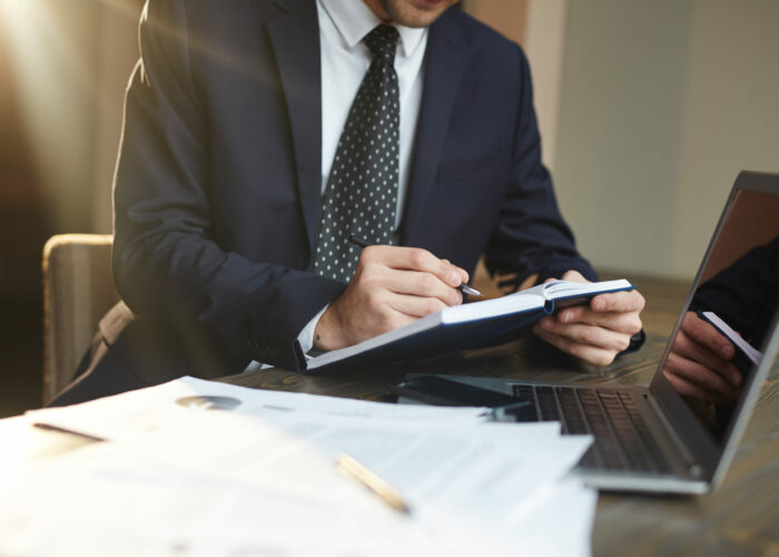 Closeup portrait of unrecognizable successful businessman wearing black formal suit  writing in planner while working with documentation and laptop